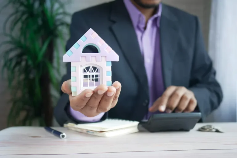 Property manager in suit holding a house model while using a calculator at a desk