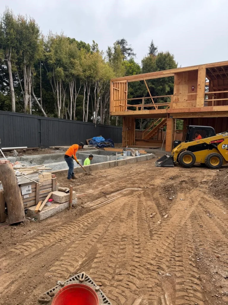Construction workers clearing debris and leveling dirt at a residential demolition and site preparation project, featuring a yellow CAT skid steer and an exposed wooden building frame.