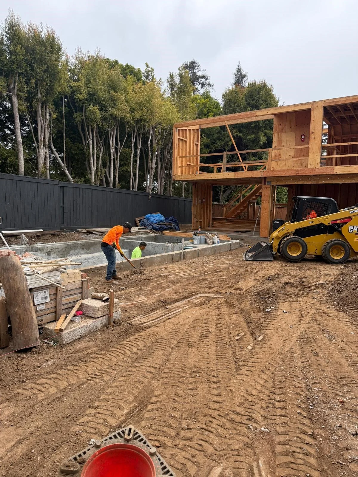Construction workers clearing debris and leveling dirt at a residential demolition and site preparation project, featuring a yellow CAT skid steer and an exposed wooden building frame.